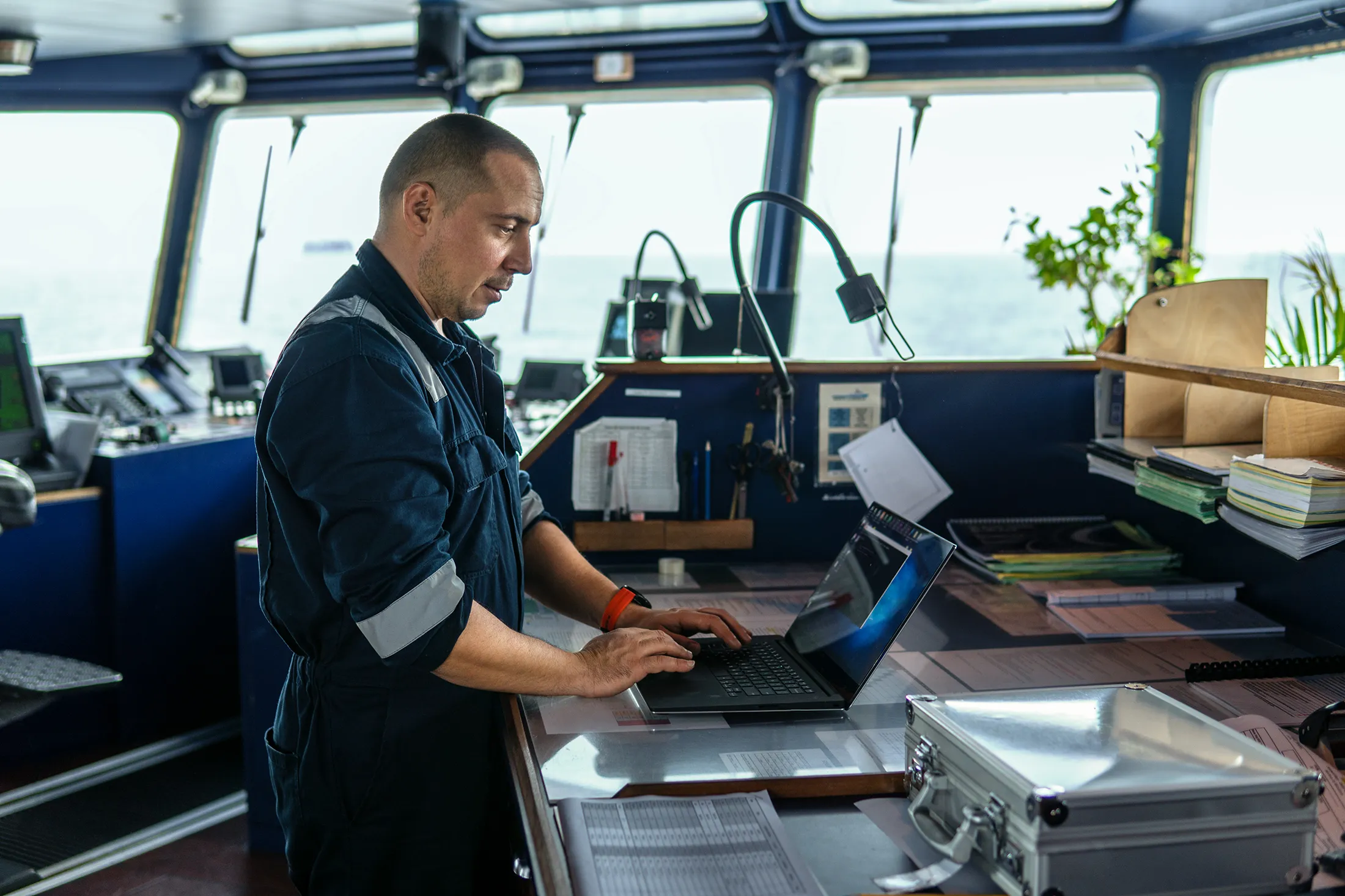 Marine navigational officer standing at a ship’s bridge, working on a laptop surrounded by charts, documents, and navigation equipment with ocean view in background.