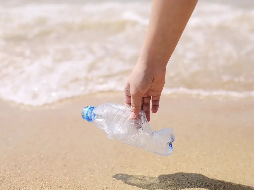 Hand picking up a crushed plastic bottle from the beach, symbolizing ocean conservation, plastic pollution awareness, and volunteer environmental cleanup efforts.