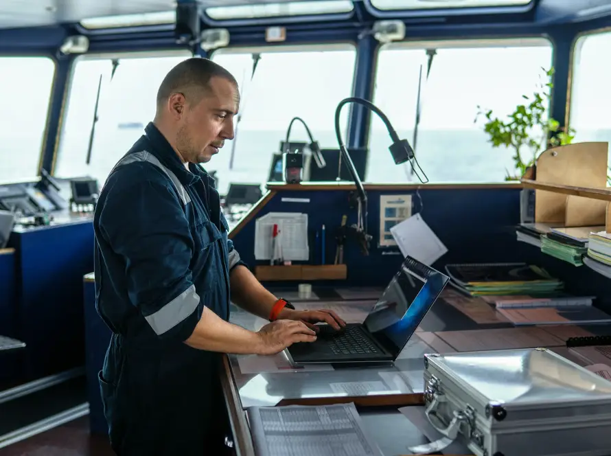 Marine navigational officer standing at a ship’s bridge, working on a laptop surrounded by charts, documents, and navigation equipment with ocean view in background.