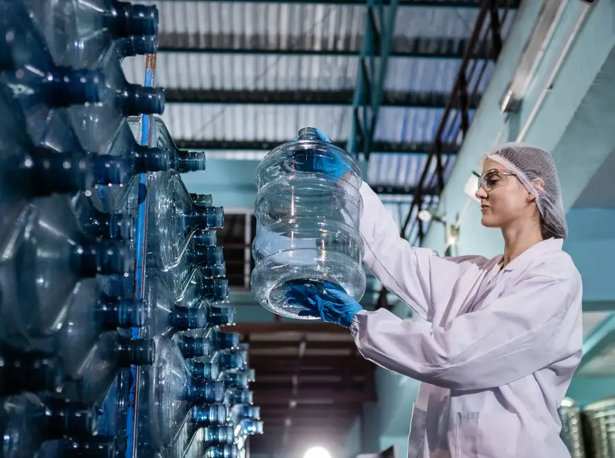 Female worker in protective gear inspecting large water bottles at a bottling facility.