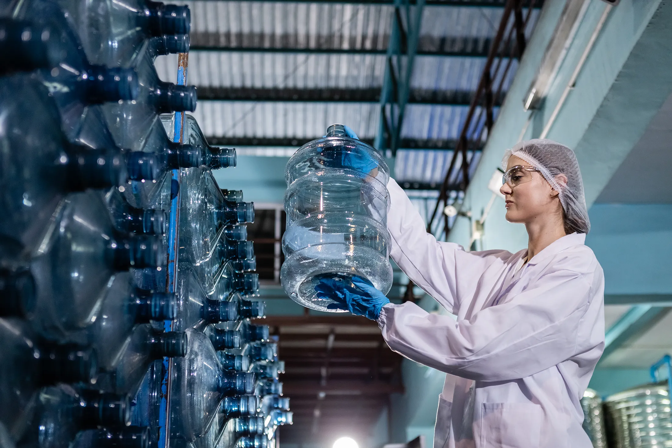 Female worker in protective gear inspecting large water bottles at a bottling facility.