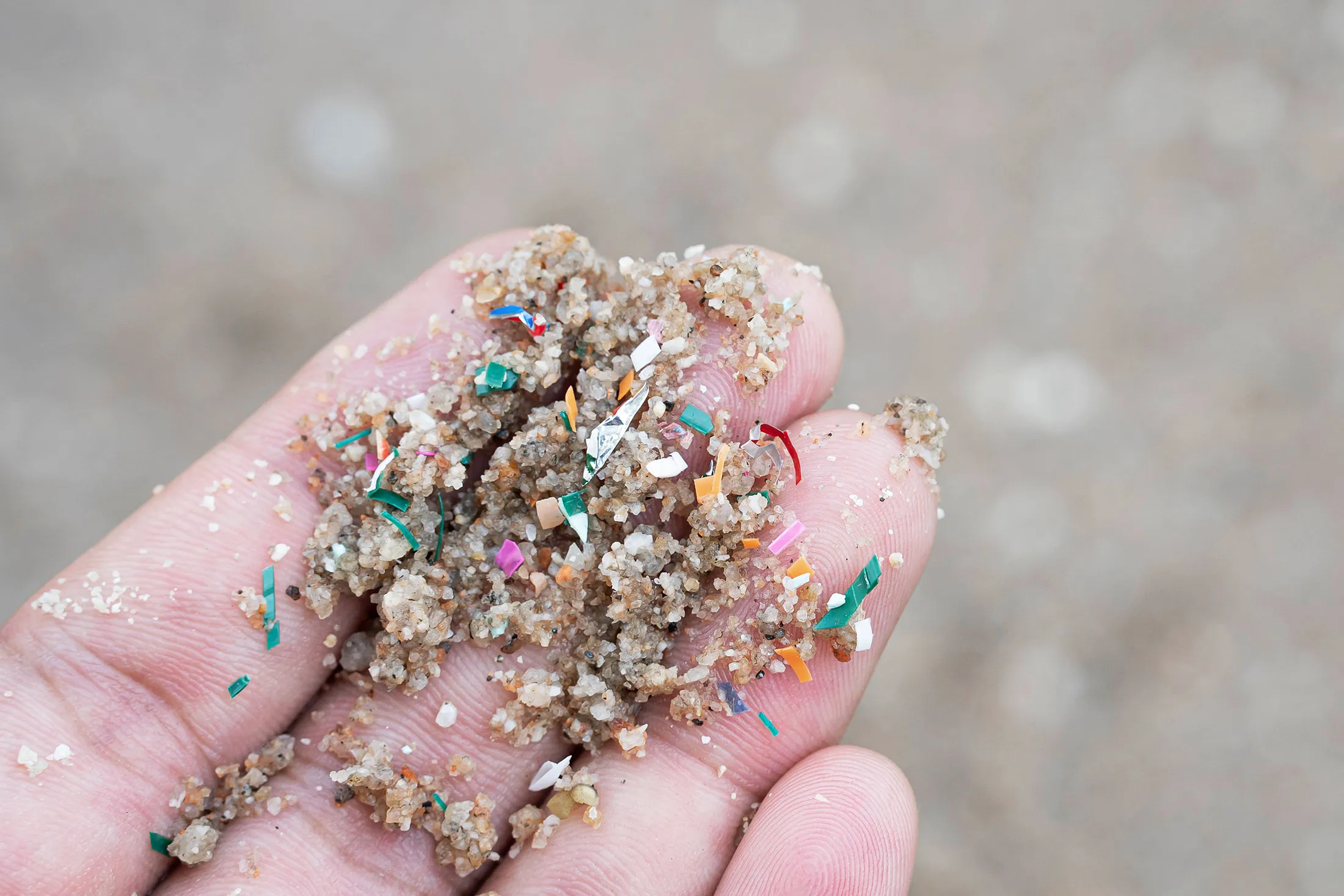 Close-up of a hand holding sand mixed with colorful microplastic particles on a beach.