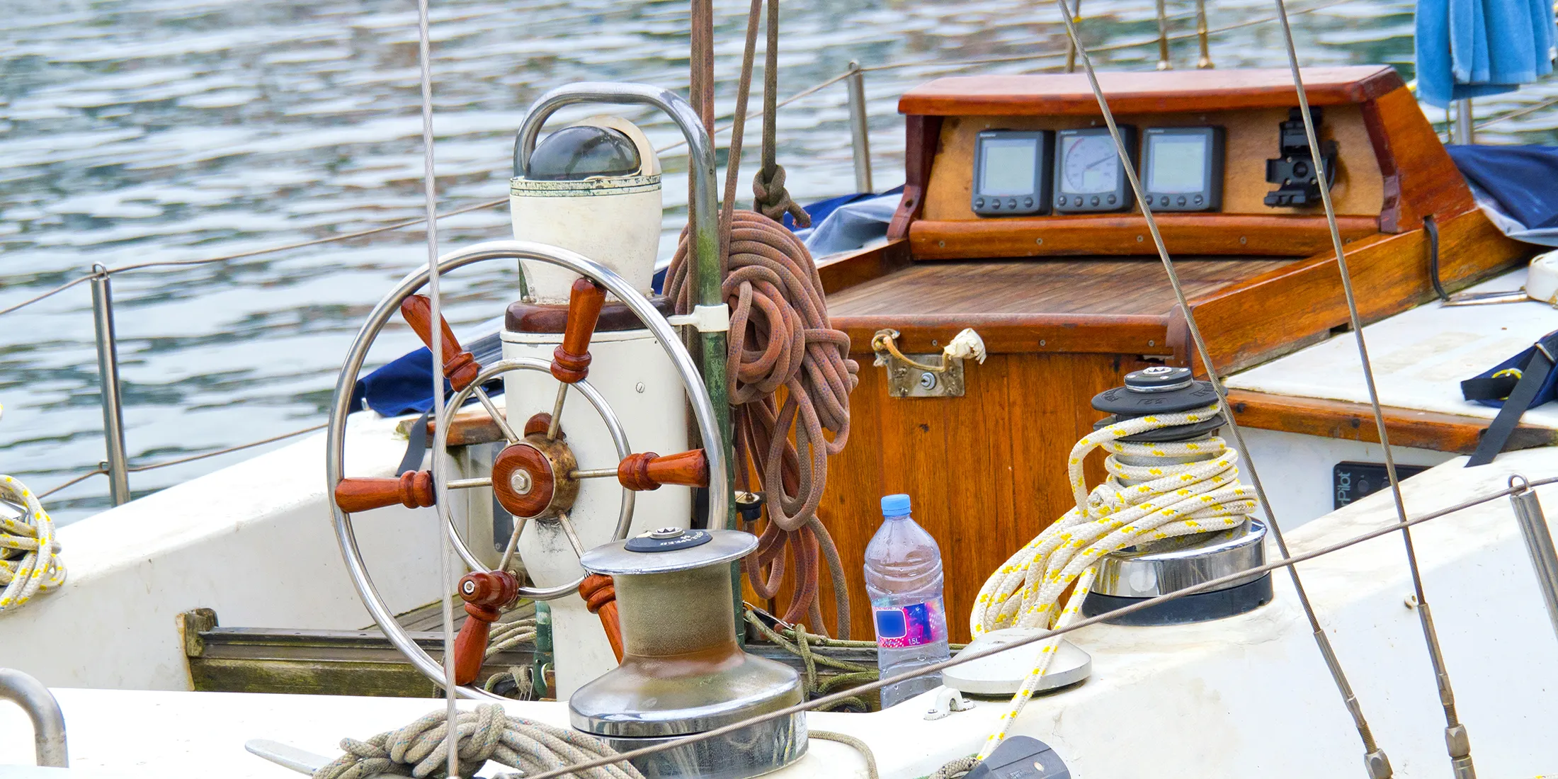 Close-up of a sailboat deck with wooden steering wheel, ropes, and navigation instruments.