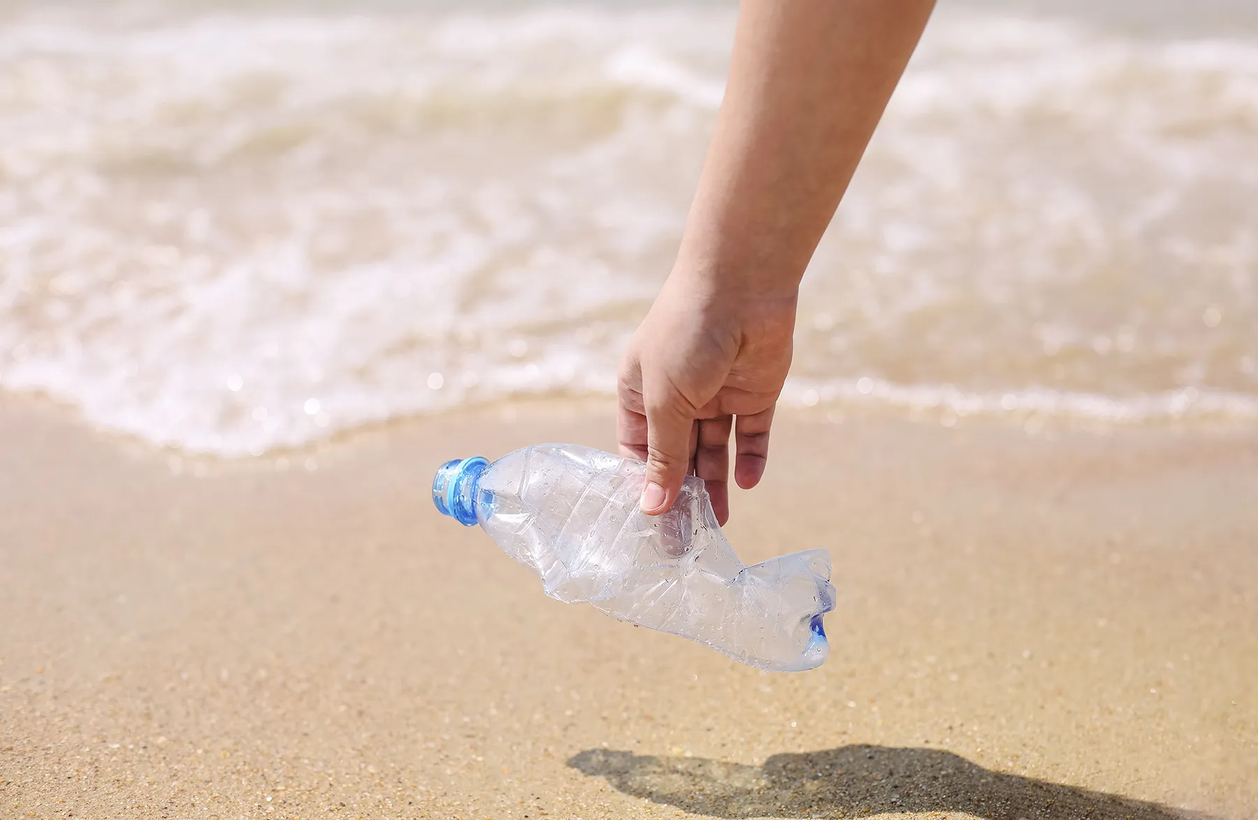 Hand picking up a crushed plastic bottle from the beach, symbolizing ocean conservation, plastic pollution awareness, and volunteer environmental cleanup efforts.