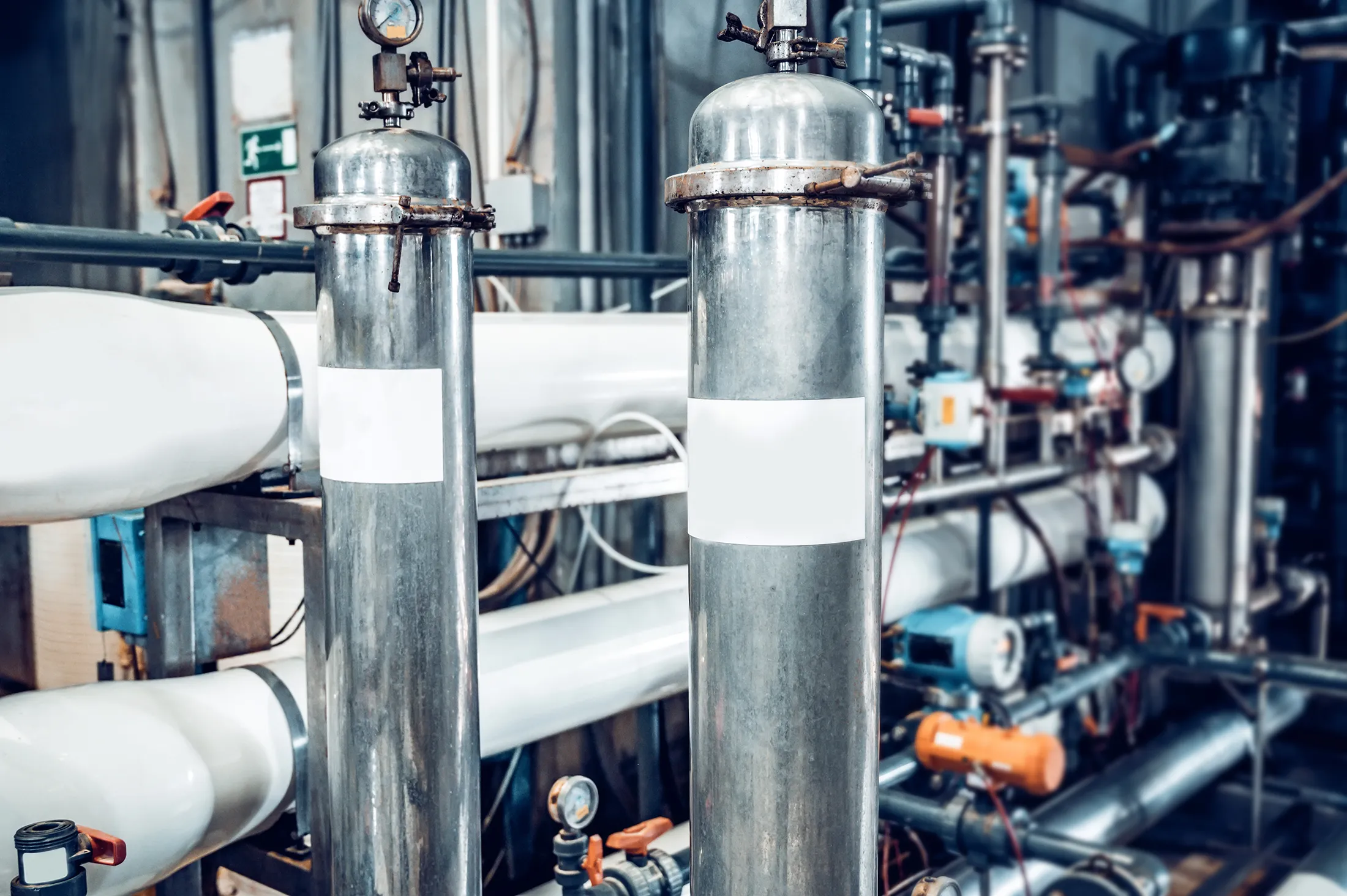 Industrial stainless steel filtration tanks and piping system inside a water purification plant, featuring gauges, valves, and connected reverse osmosis equipment.