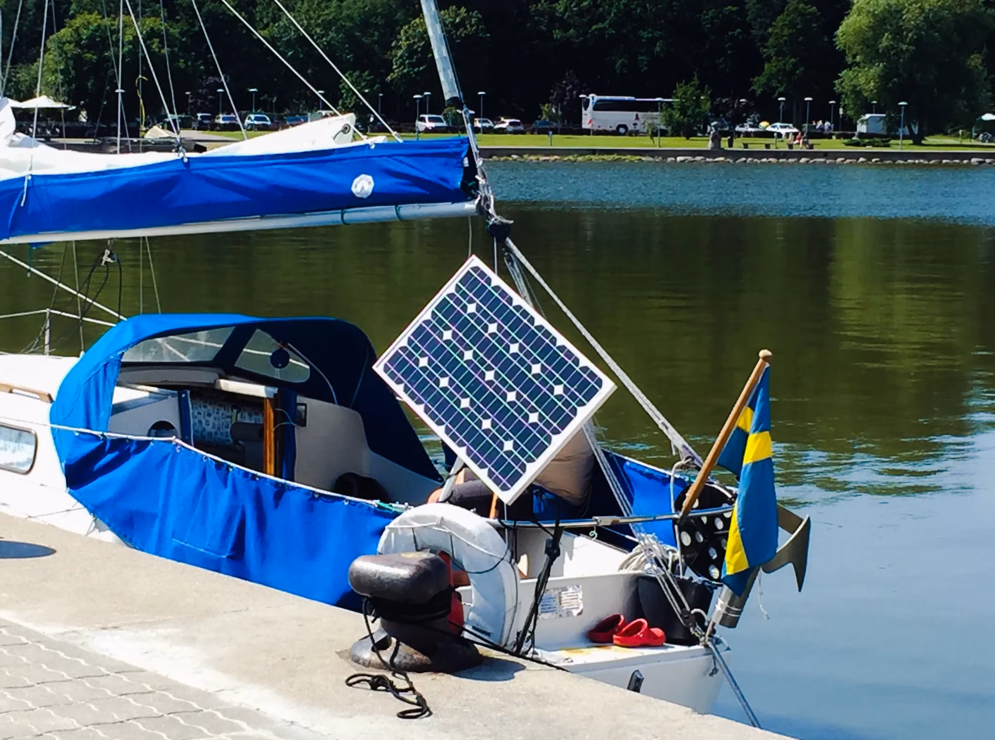 Sailboat docked on calm water with a mounted solar panel and blue canvas covers on a sunny day.